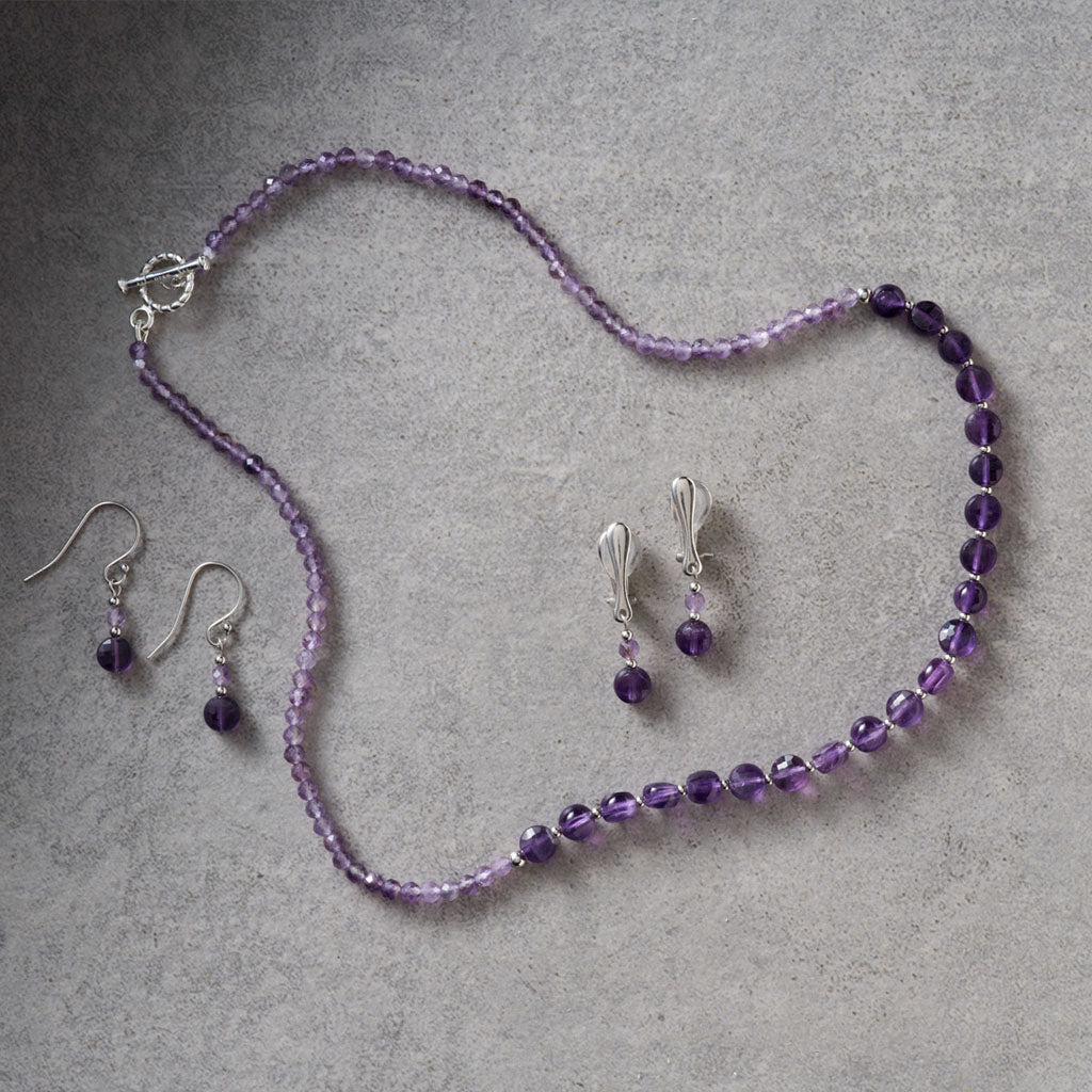 A jewelry set featuring a beaded necklace and matching drop earrings in shades of purple, accented with silver-tone metal findings, displayed on a textured gray background.