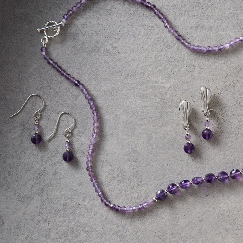 A jewellery set featuring a beaded necklace and matching drop earrings in purple beads with silver-toned metal findings, arranged on a textured gray surface.