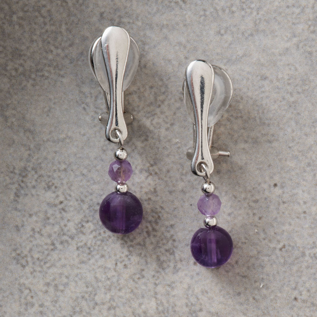 A pair of silver-toned clip-on earrings with faceted purple beads, displayed on a textured gray surface.