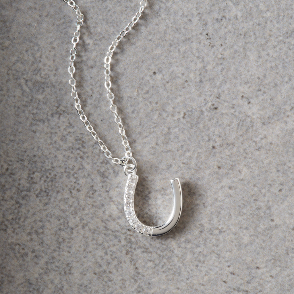 A silver horseshoe pendant necklace with a row of clear crystals along the left side, resting on a textured gray surface.