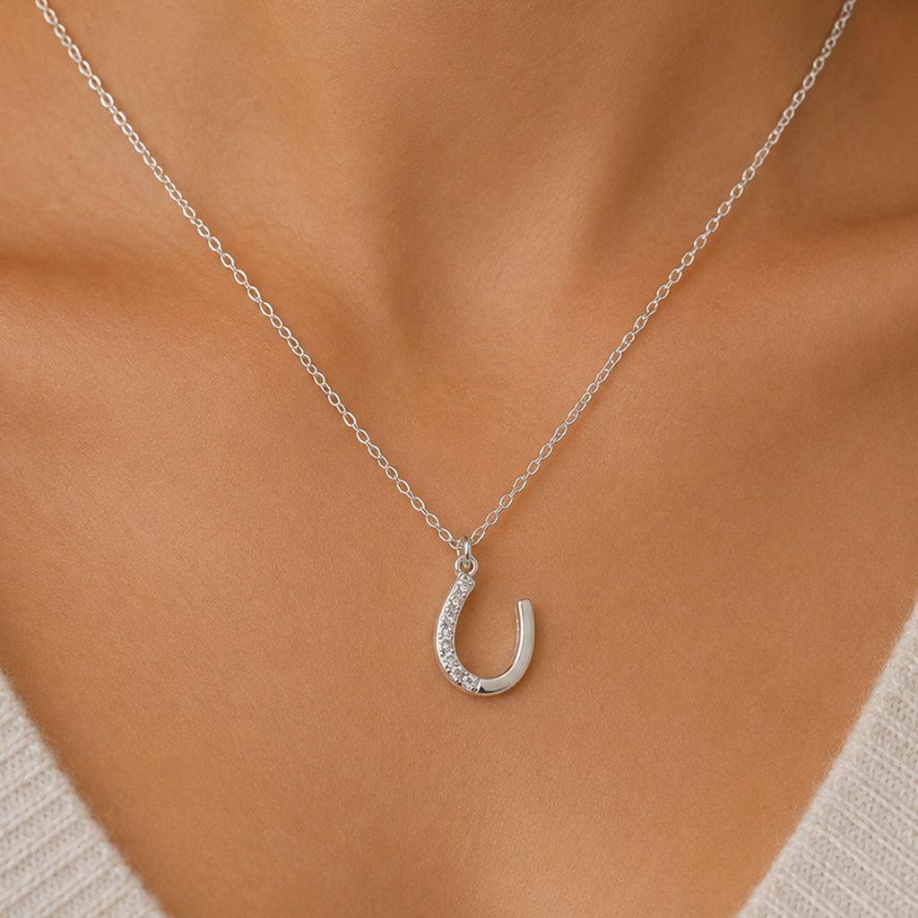 A silver horseshoe pendant necklace with small clear stones along the lower curve, worn on a model's neck over a light-colored knit top.
