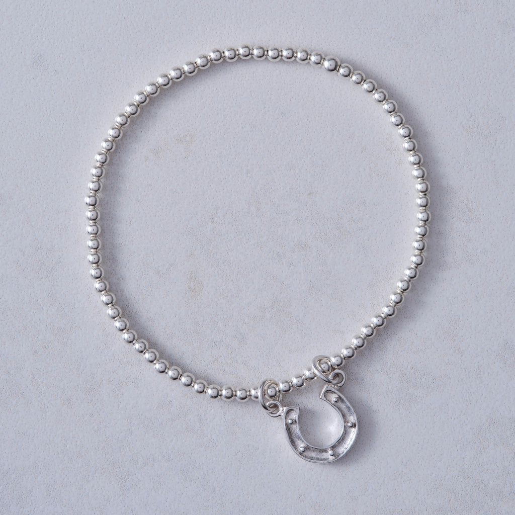 A silver beaded bracelet with a small silver horseshoe charm, displayed on a light gray textured background.