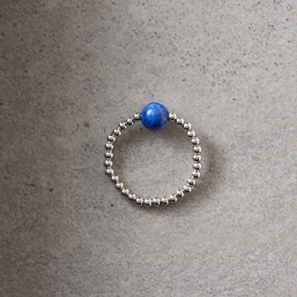 A silver beaded ring with a single round blue bead at the top, photographed on a textured gray surface.
