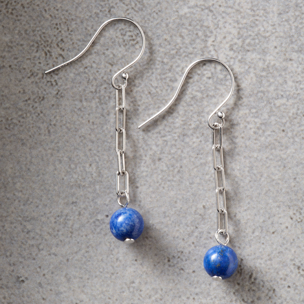 A pair of silver dangle earrings with paperclip chain links and round blue lapis lazuli beads, displayed on a textured gray surface.