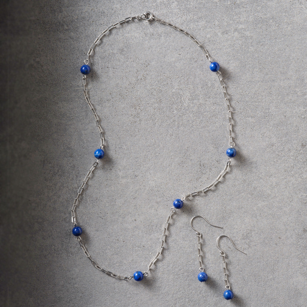A silver-toned chain necklace and matching drop earrings with small round blue beads, arranged on a textured gray surface.