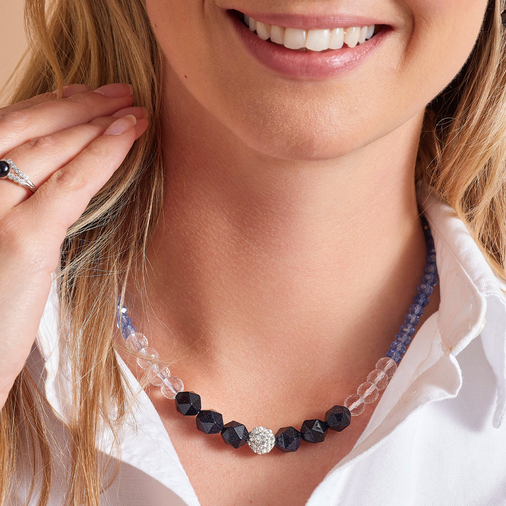 A close-up lifestyle shot of a woman wearing a beaded necklace. The necklace features faceted black beads, clear crystal beads, and a central round silver pavé cluster, with a blue beaded cord visible behind it.