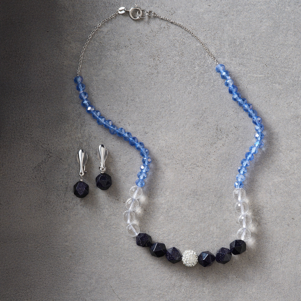 A beaded necklace and matching drop earrings laid flat on a textured gray surface. The necklace has faceted light blue beads, clear beads, dark navy faceted beads, and a central silver-toned round bead with clear stones.