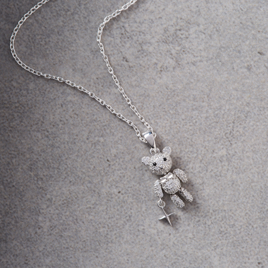 A silver necklace featuring a teddy bear pendant covered in small clear stones, holding a small silver cross charm, displayed on a textured gray surface.