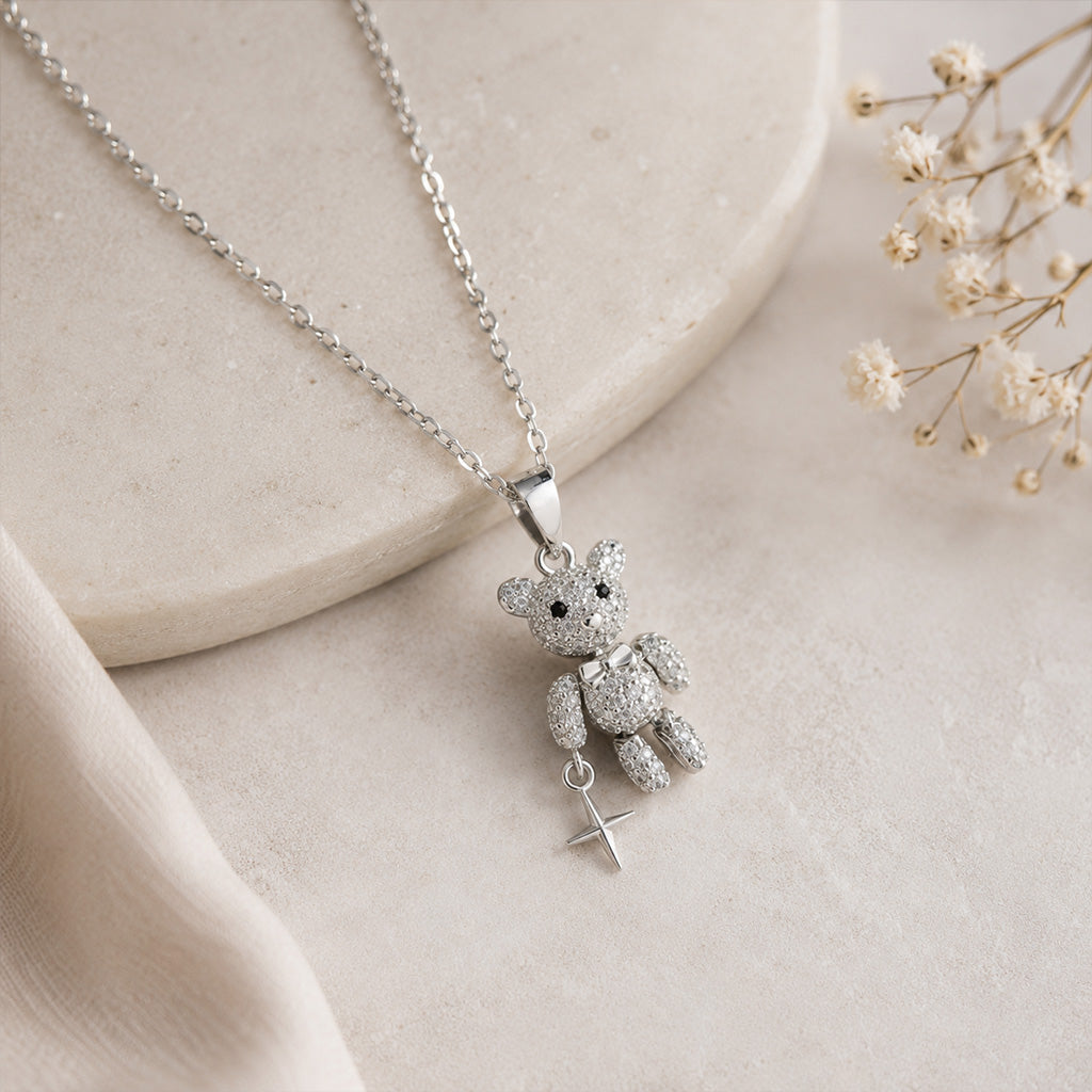 A silver necklace with a teddy bear pendant and a small cross charm, displayed on a light beige stone slab with dried baby's breath flowers.