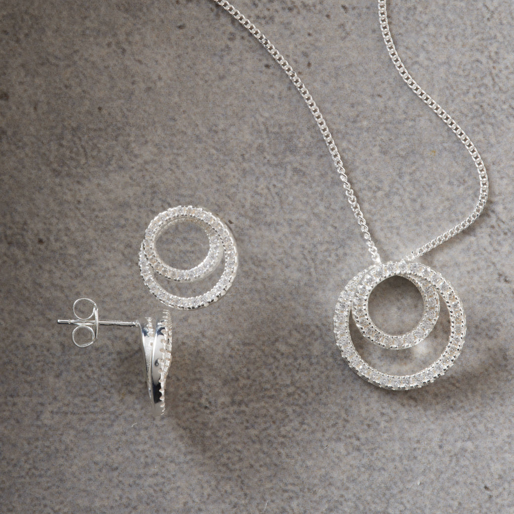 Silver-tone jewelry set featuring a necklace with a double-ring pendant and matching stud earrings, displayed on a textured gray surface.