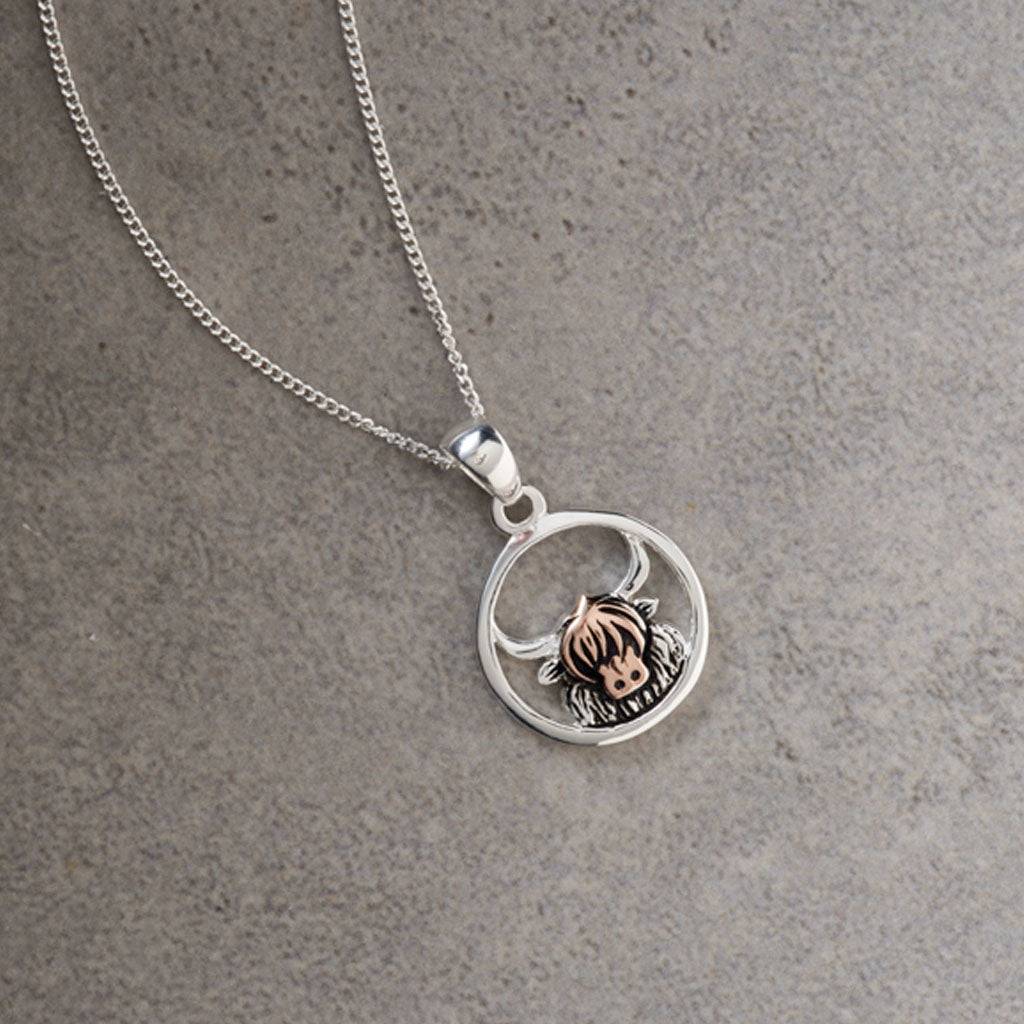 A silver chain necklace with a circular pendant featuring a small cow design in rose gold and silver, displayed on a textured gray surface.