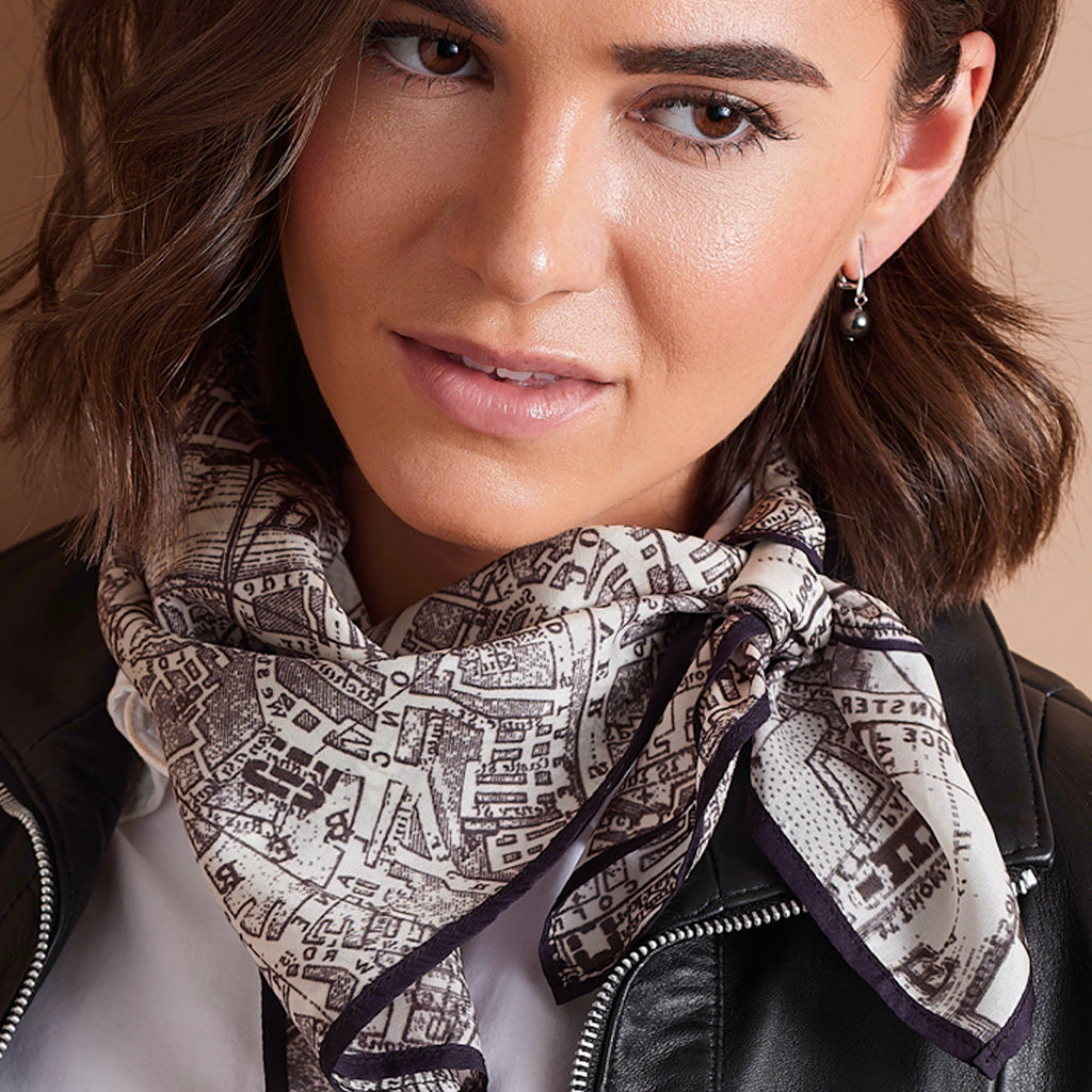 A close-up of a woman wearing a black leather jacket and a cream scarf with a dark brown map-like print and dark purple border.