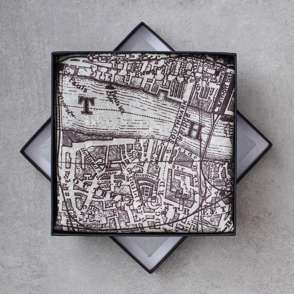 A square scarf with a black and white vintage map print, presented in a black box with a geometric star-shaped base on a gray textured background.