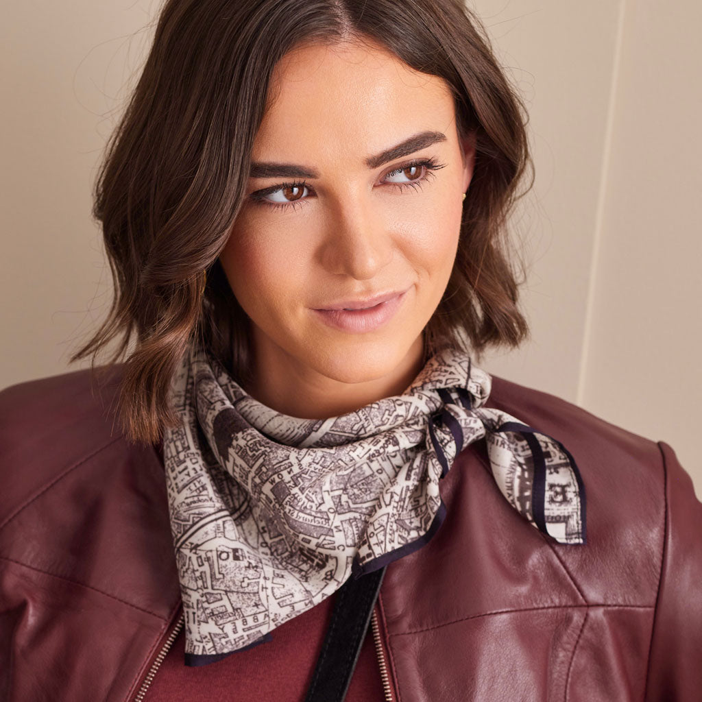 A woman with shoulder-length brown hair wearing a burgundy leather jacket and a patterned scarf. The scarf features a black and beige map-like print with a dark border, styled around her neck.