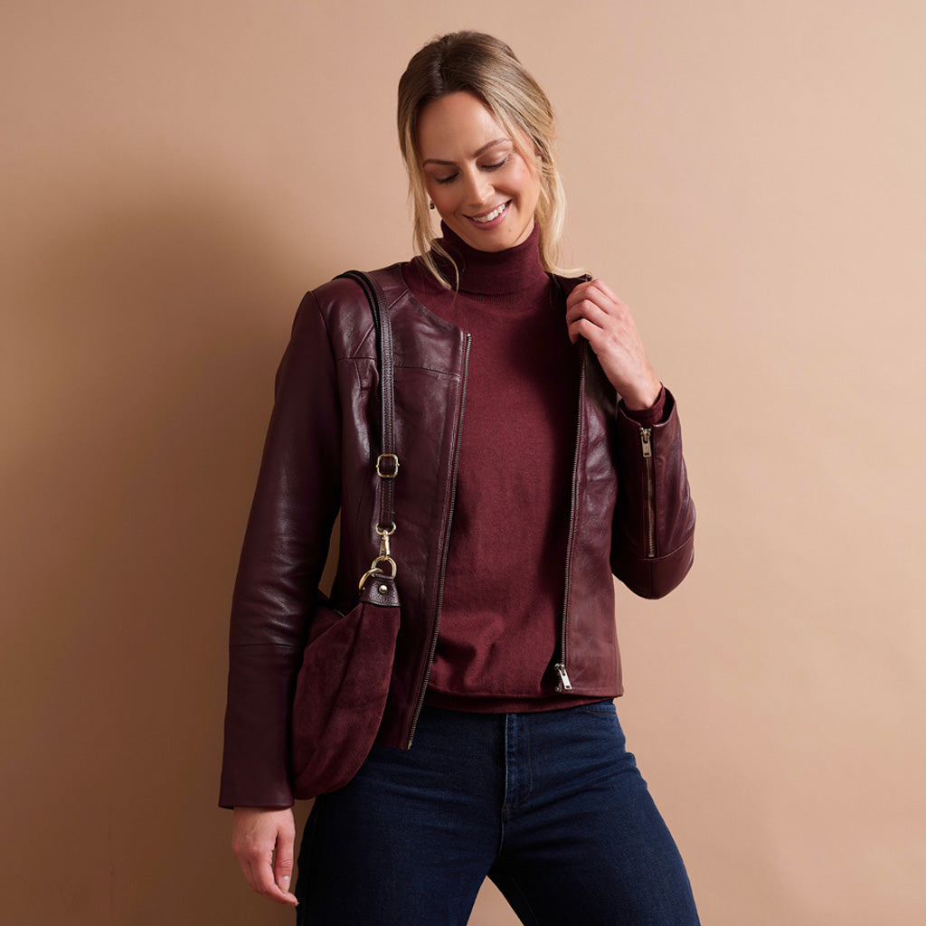 A woman wearing a dark burgundy leather moto jacket over a matching burgundy turtleneck sweater and dark blue jeans, posing against a solid beige background.
