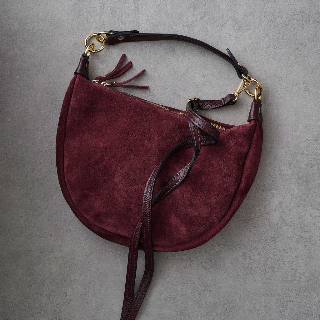 A burgundy suede crescent-shaped shoulder bag with dark brown leather straps and gold-tone hardware, laid flat on a textured gray background.