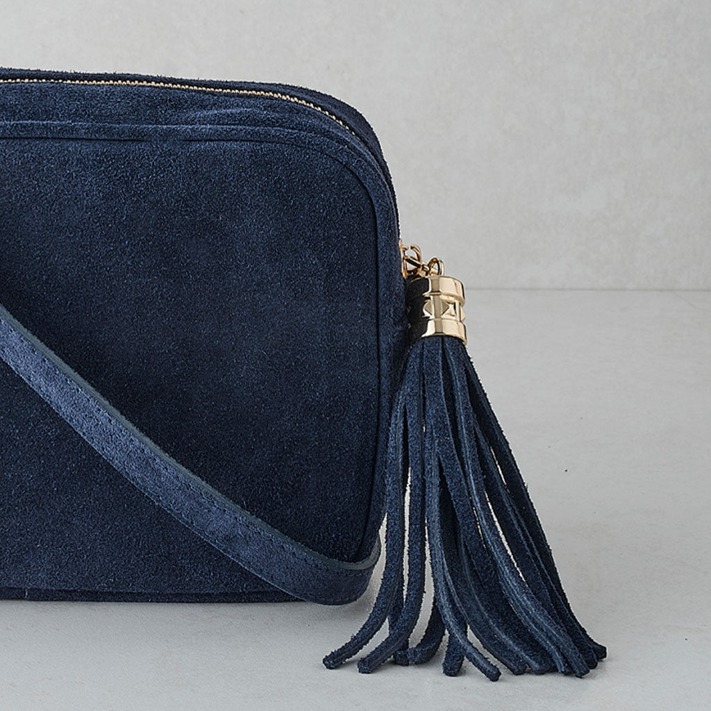 A navy blue suede crossbody bag with a matching shoulder strap and a decorative tassel charm featuring a gold-tone metal cap, shown against a neutral light gray background.