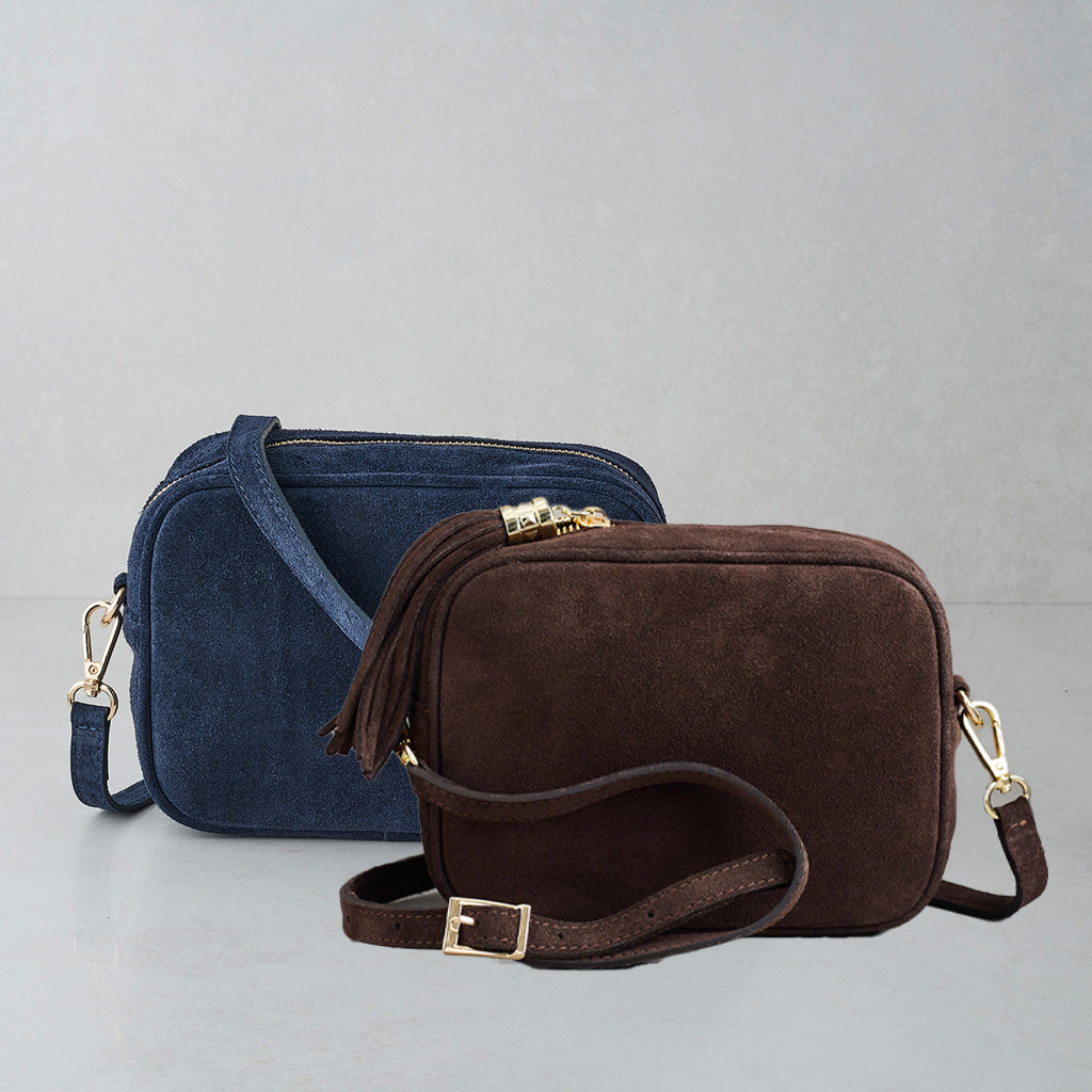 Two small crossbody bags with rectangular shapes and adjustable straps, shown on a light gray studio background. The front bag is dark brown suede with a tassel detail and gold-tone hardware, and the bag behind it is blue suede with matching blue strap and hardware.