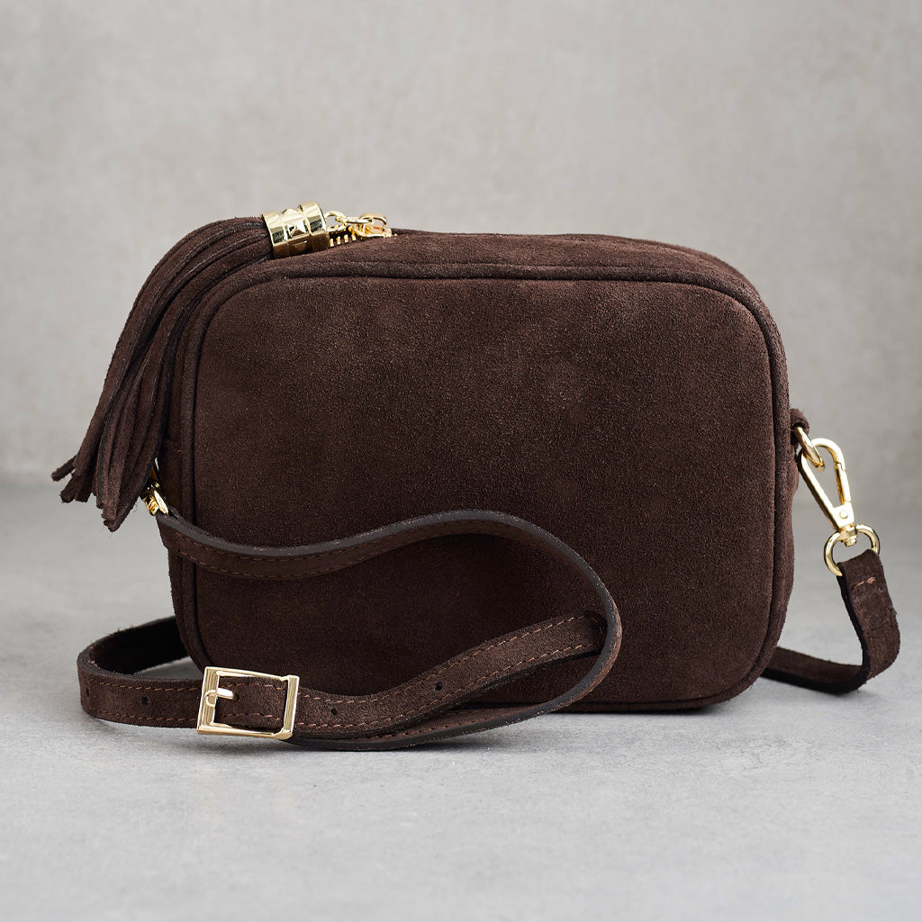 A dark brown suede crossbody camera bag with a tassel zipper pull, adjustable shoulder strap, and gold-tone hardware, shown on a neutral gray background.