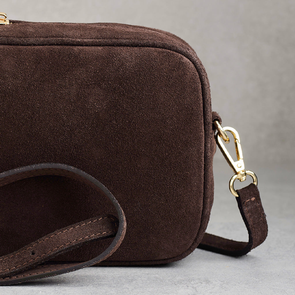 A close-up of a dark brown suede crossbody bag with a matching leather strap and gold-tone hardware, set against a neutral gray background.