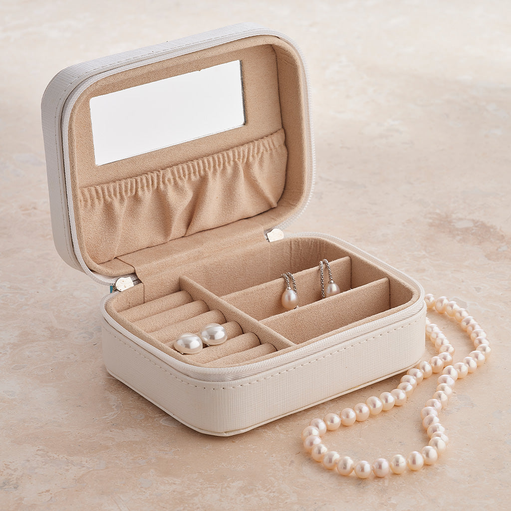 An open cream-colored faux leather jewelry organizer box with a mirror in the lid, pearl necklace, and pearl earrings displayed inside on a beige stone surface.