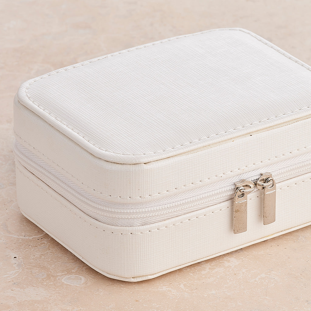 A cream-colored double zipper jewelry box with a textured fabric finish and gold-tone zipper pulls, resting on a light beige stone surface.