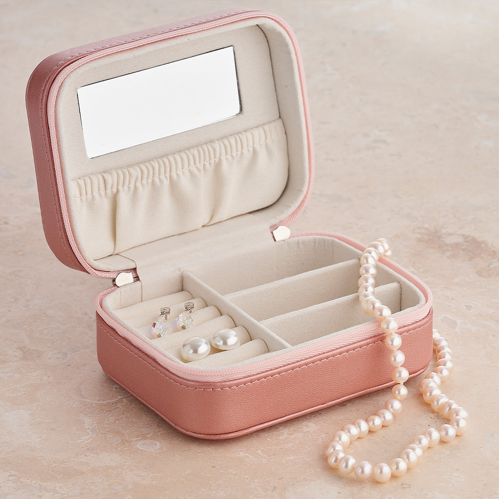 An open pink faux-leather jewelry organizer box with a mirror on the inside lid, pearl earrings, and a pearl necklace resting on the front, set on a beige stone-like surface.