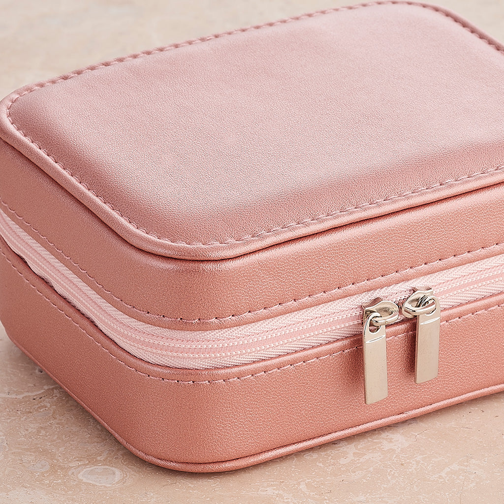 A compact rectangular jewelry box in dusty pink faux leather with a double zipper closure and silver-toned metal pulls, shown on a light beige textured surface.