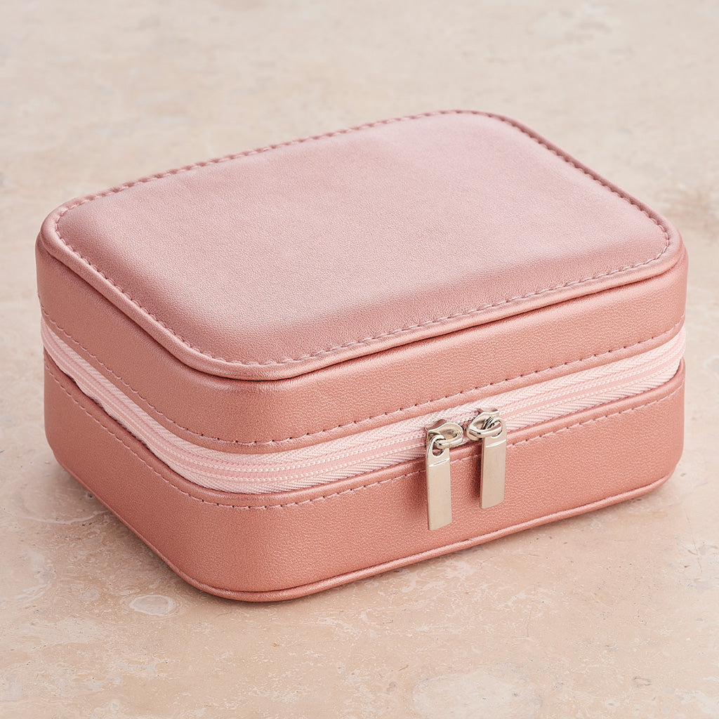 A compact, rectangular pink faux-leather jewelry box with a double zipper closure and silver-toned metal pulls, set against a neutral beige textured background.