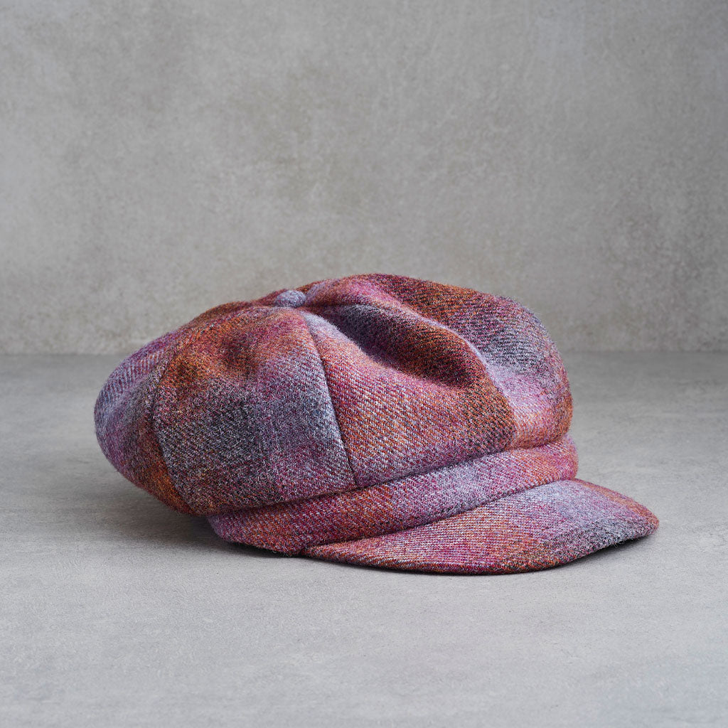 A vintage-style newsboy cap in a textured tweed fabric with a mottled pattern of purple, pink, and brown, resting on a gray concrete surface.