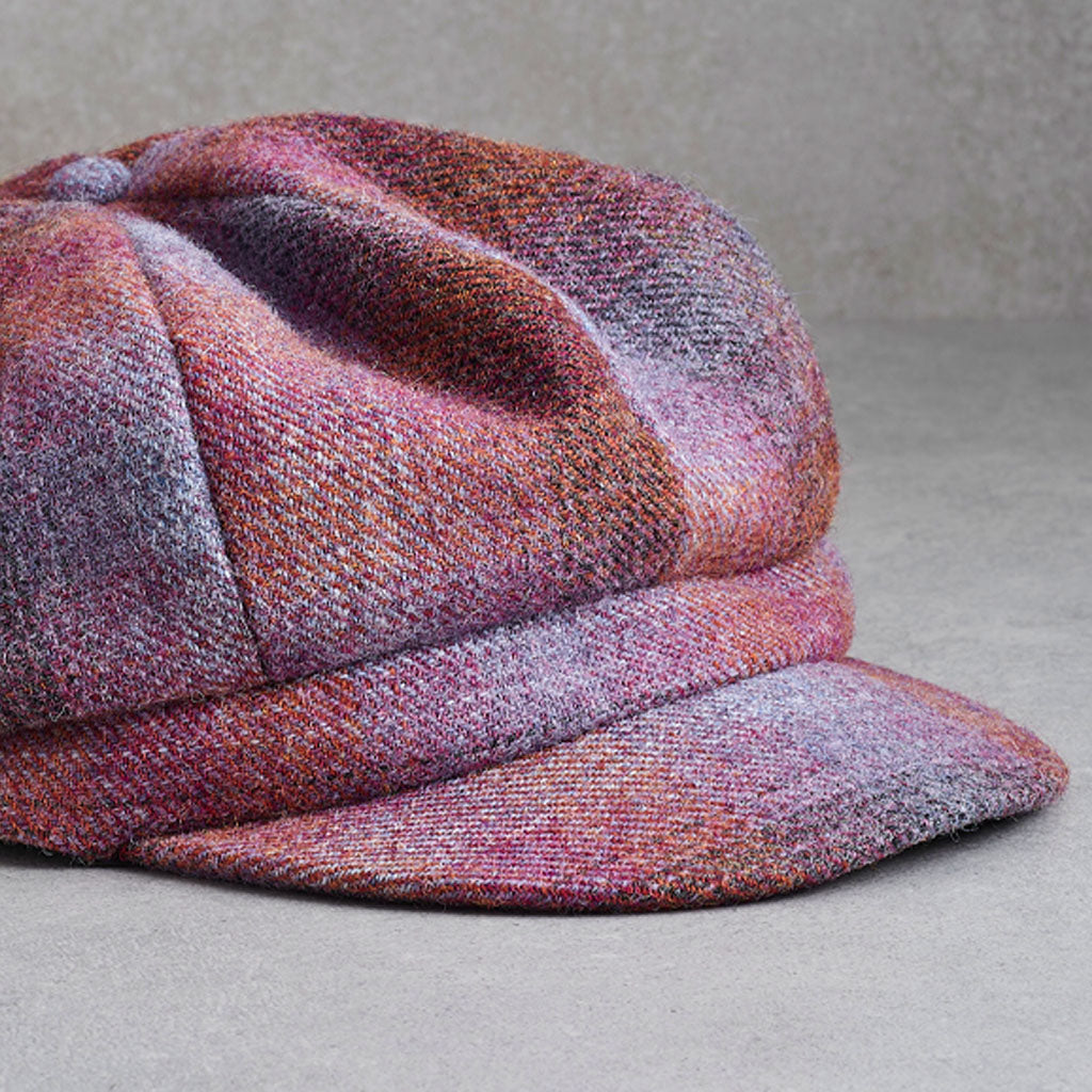 A close-up of a textured tweed flat cap in a heathered blend of pink, purple, and brown tones, resting on a neutral gray surface.