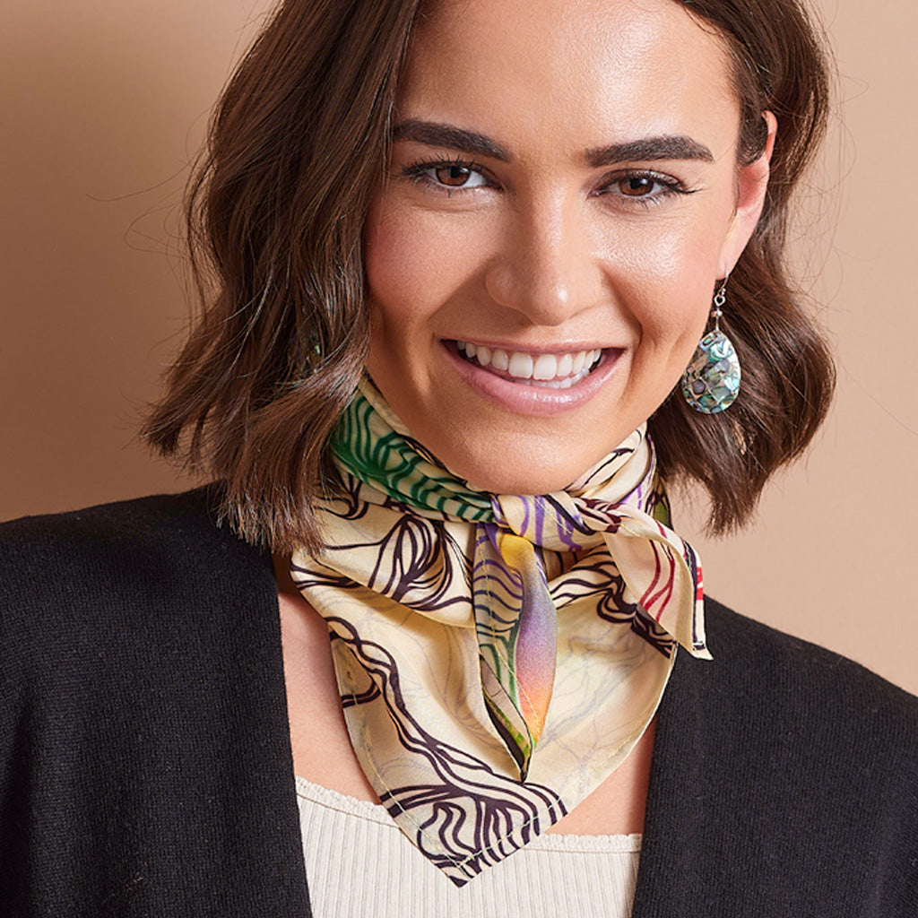 A woman with short brown hair smiling, wearing a beige silk scarf with a colorful botanical and abstract pattern, styled with a black cardigan and a cream top against a solid tan background.