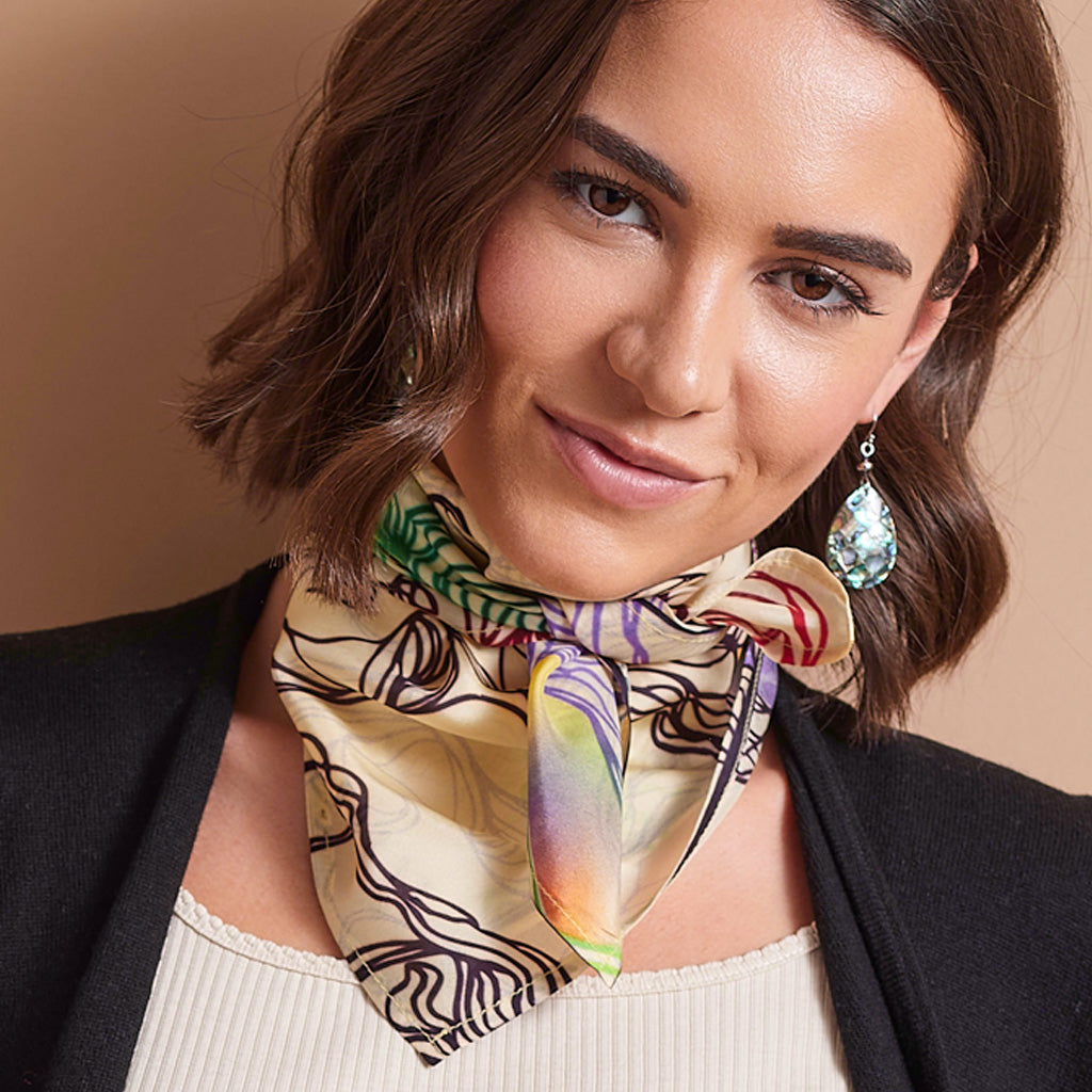 A woman with short brown hair models a beige silk scarf with black abstract line patterns and colorful gradient accents, paired with a black blazer and cream top against a warm beige background.