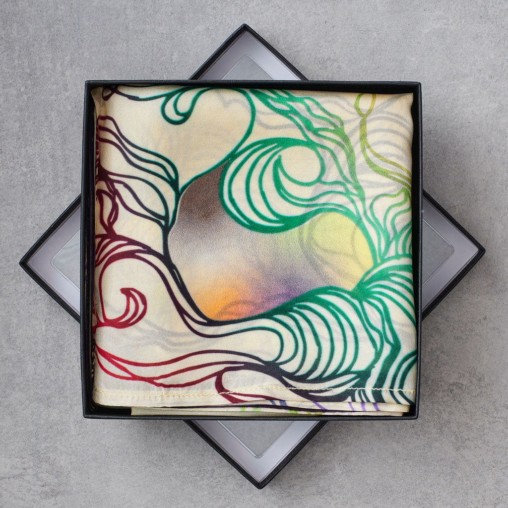 A square silk scarf with an abstract wave pattern in green, red, yellow, and purple, presented in a black gift box on a gray textured background.