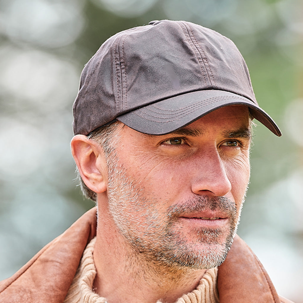 A close-up portrait of a man wearing a dark gray baseball cap and a brown jacket, set against a soft, blurred outdoor background.