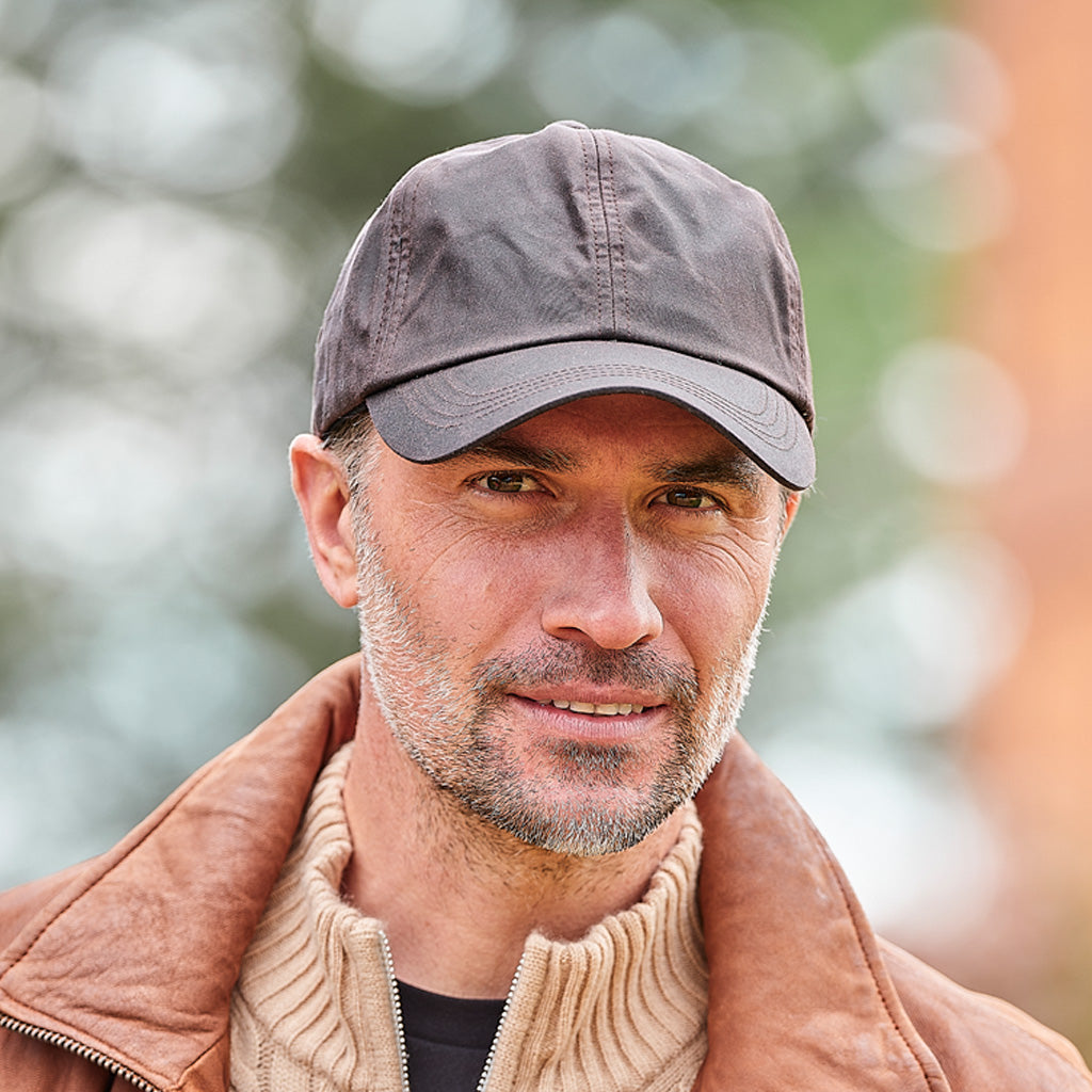 A close-up portrait of a man wearing a dark gray baseball cap and a brown leather jacket over a beige ribbed sweater, set against a soft, blurred outdoor background.