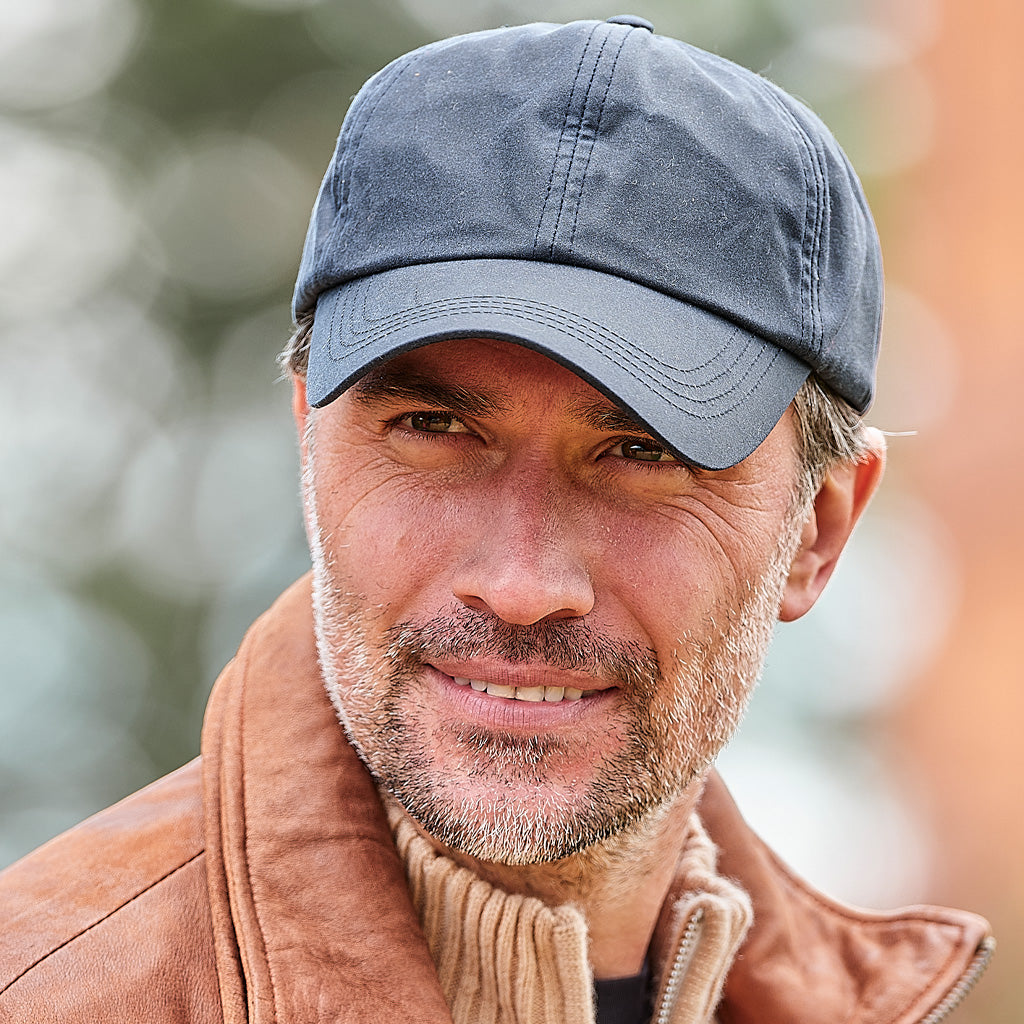 A close-up portrait of a man wearing a dark gray baseball cap and a brown leather jacket over a beige sweater, set against a soft, blurred outdoor background.