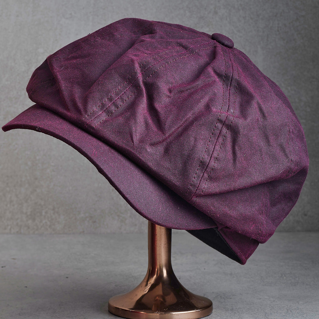 A dark purple newsboy cap with a textured fabric finish, displayed on a metallic rose gold stand against a gray background.
