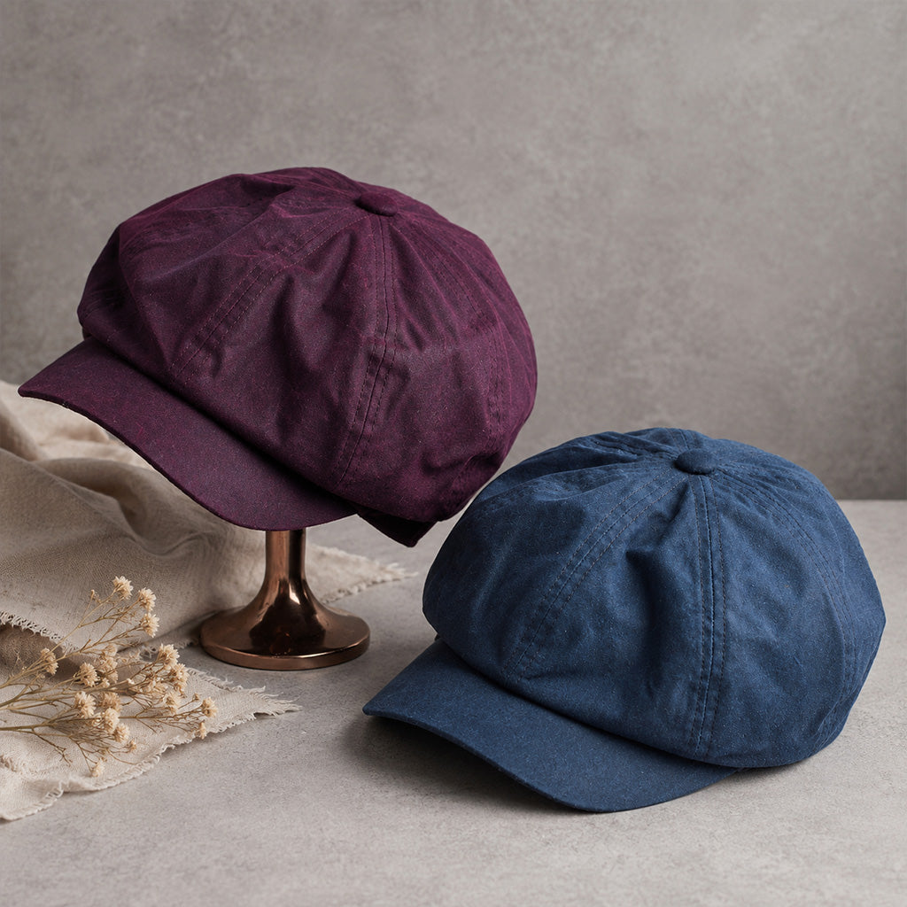 Two flat-brim newsboy caps displayed on a tabletop: one in deep purple on a small stand and one in blue resting beside it, with a neutral textured gray background and dried flowers as props.