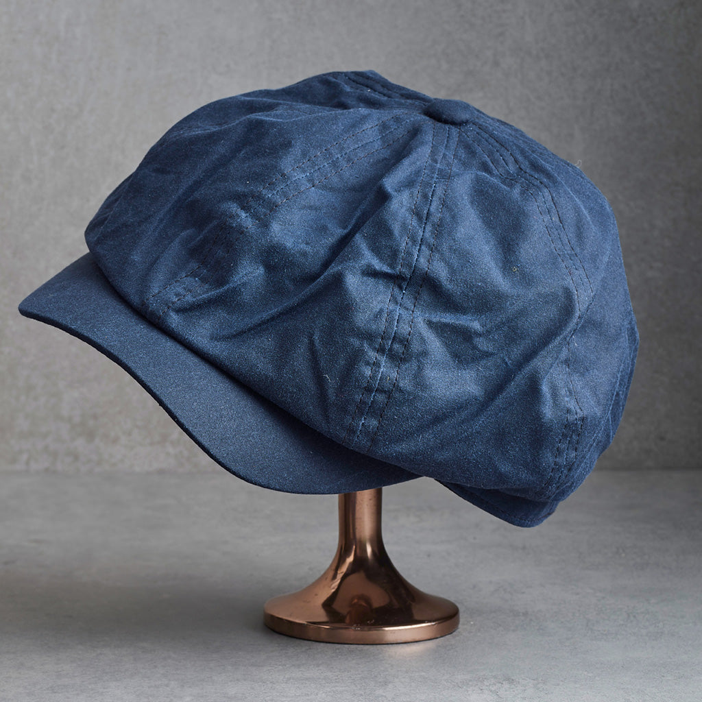 A navy newsboy cap with a textured wax fabric finish, displayed on a metallic rose gold stand against a gray background.