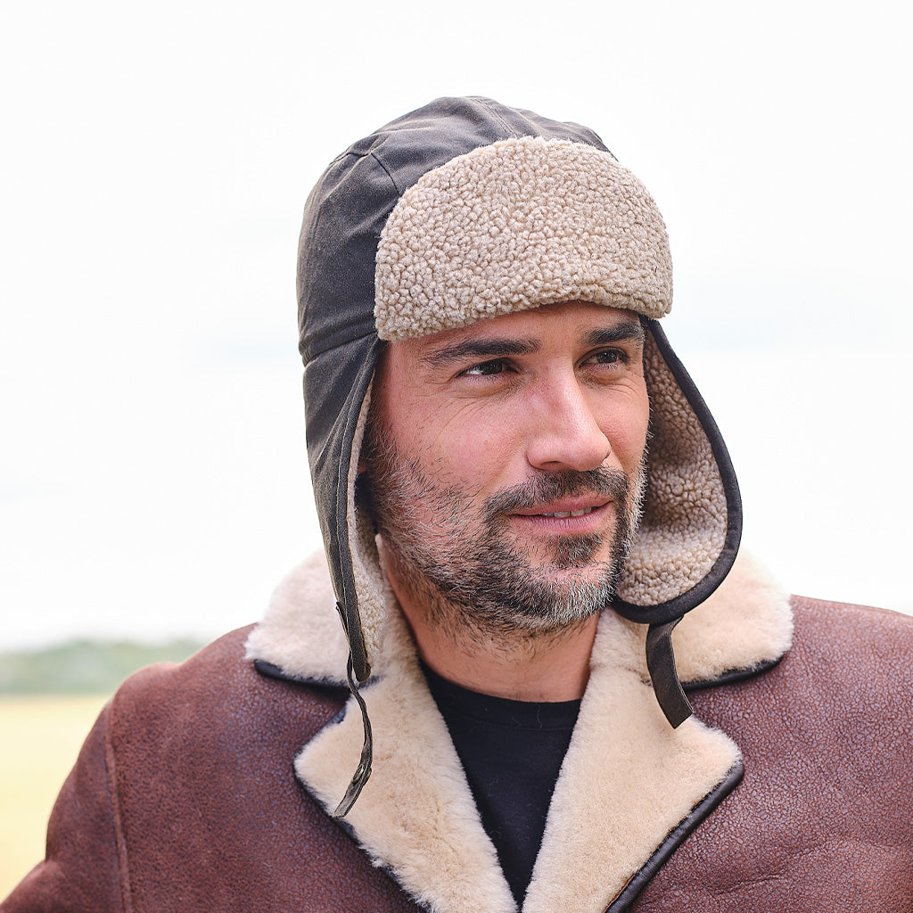 A man wearing a brown shearling-lined aviator trapper hat with ear flaps and a brown shearling-lined shearling coat, photographed outdoors against a bright, overcast sky.