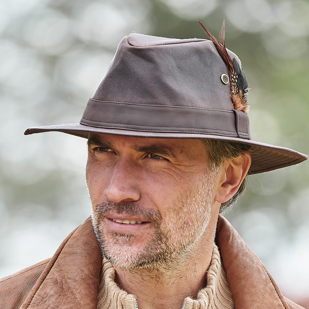 A close-up of a man wearing a brown wide-brim fedora hat with a feather accent and a brown leather jacket, set against a soft, blurred outdoor background.