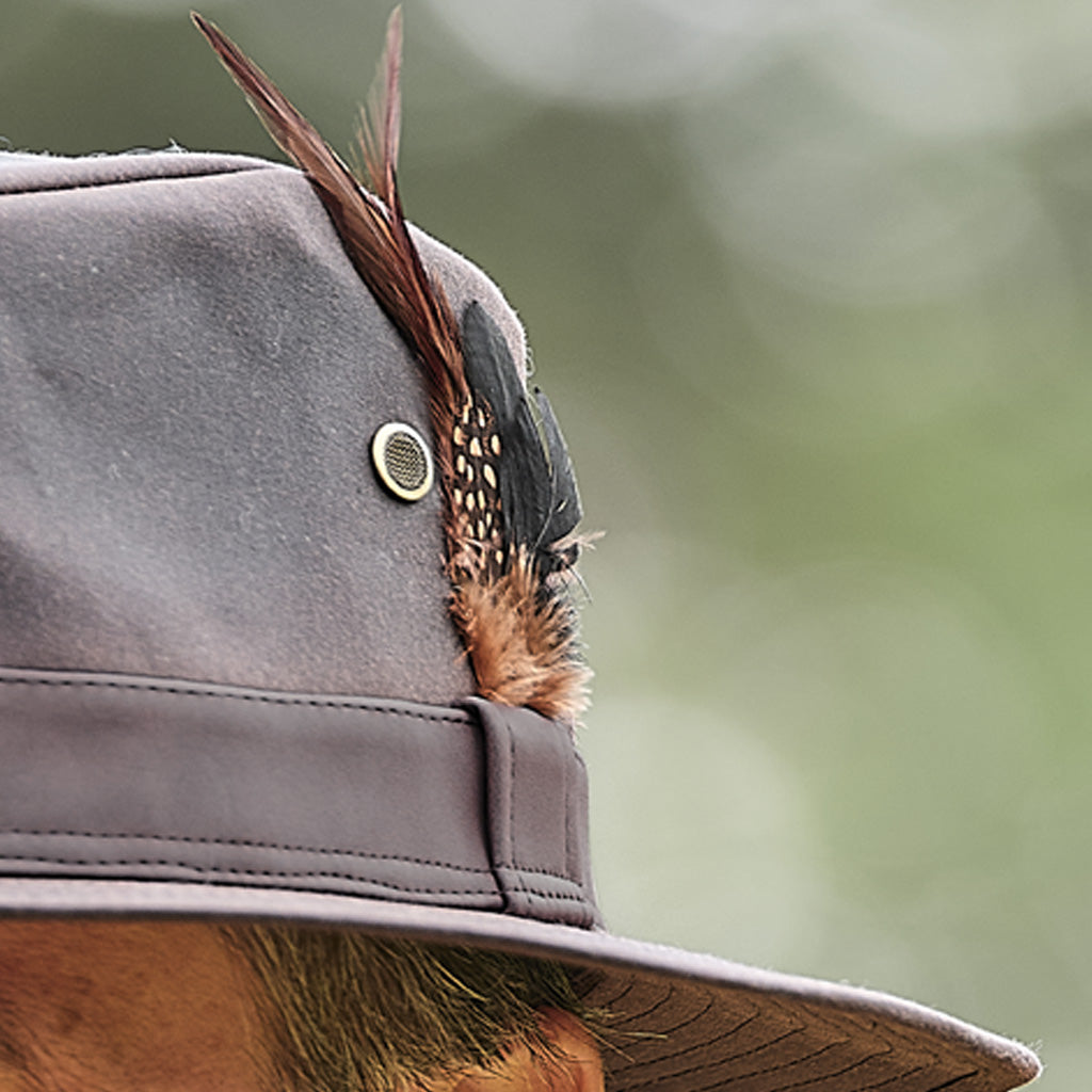 Close-up of a gray wide-brim hat with a feather accent on the side, worn by a person whose ear and hair are partially visible against a soft blurred green background.