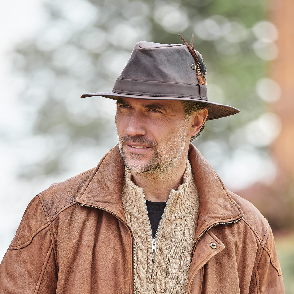 A man wearing a brown leather jacket, a beige cable-knit sweater, and a brown leather fedora hat with a feather accent, set against a soft, blurred outdoor background.