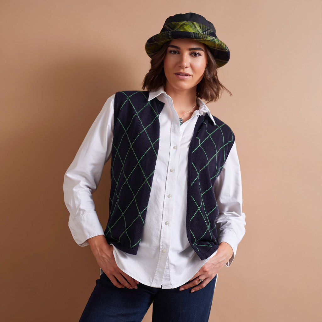 A woman modeling a sleeveless dark navy vest with a green argyle pattern, worn over a white button-down shirt and paired with a green and black patterned bucket hat, against a solid tan studio background.