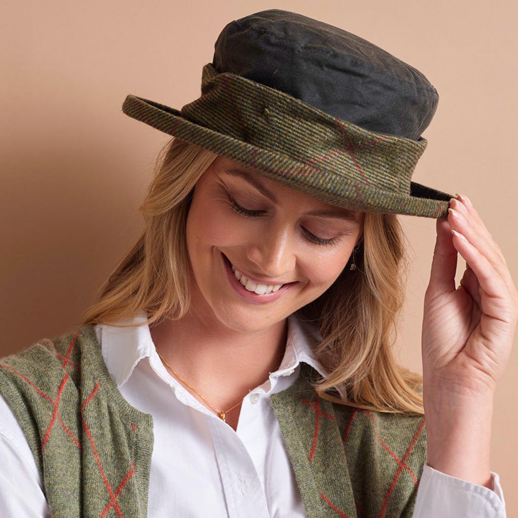 A woman wearing a vintage-style wide-brim hat in olive green tweed with a black crown, paired with a matching green argyle sweater vest over a white collared shirt, set against a solid warm beige background.