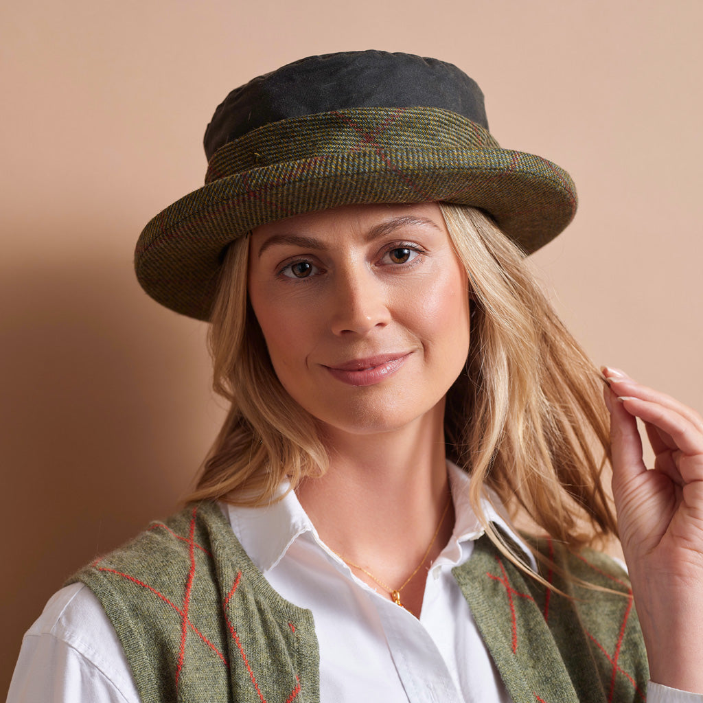 A woman wearing a textured olive green and black wide-brim hat, styled with a white collared shirt and an olive green knit vest with red lines, set against a solid beige background.