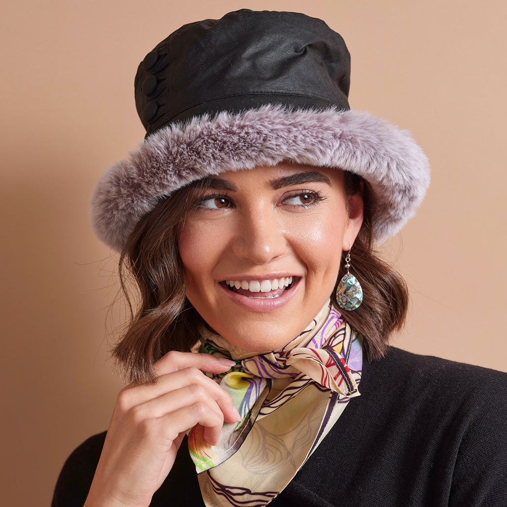 A smiling woman wearing a black bucket hat with a light gray faux fur brim, styled with a patterned silk neck scarf and a black top against a solid beige background.