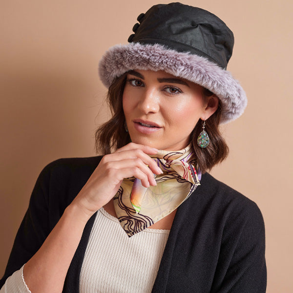 A woman wearing a black bucket hat with a light purple faux-fur brim, a cream ribbed top, a black cardigan, and a patterned scarf, posed against a solid beige background.