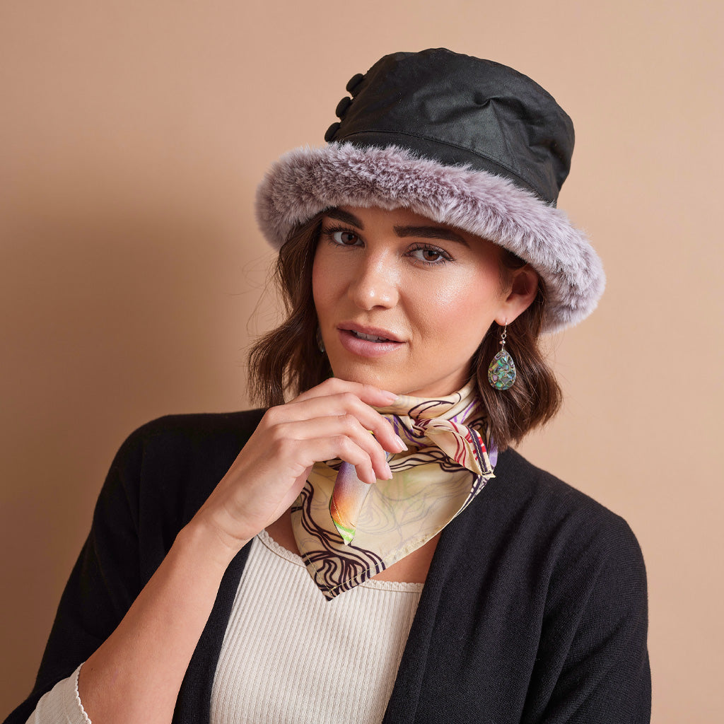 A woman wearing a black bucket hat with a light purple faux-fur brim, a cream ribbed top, a black cardigan, and a patterned scarf, set against a solid beige background.